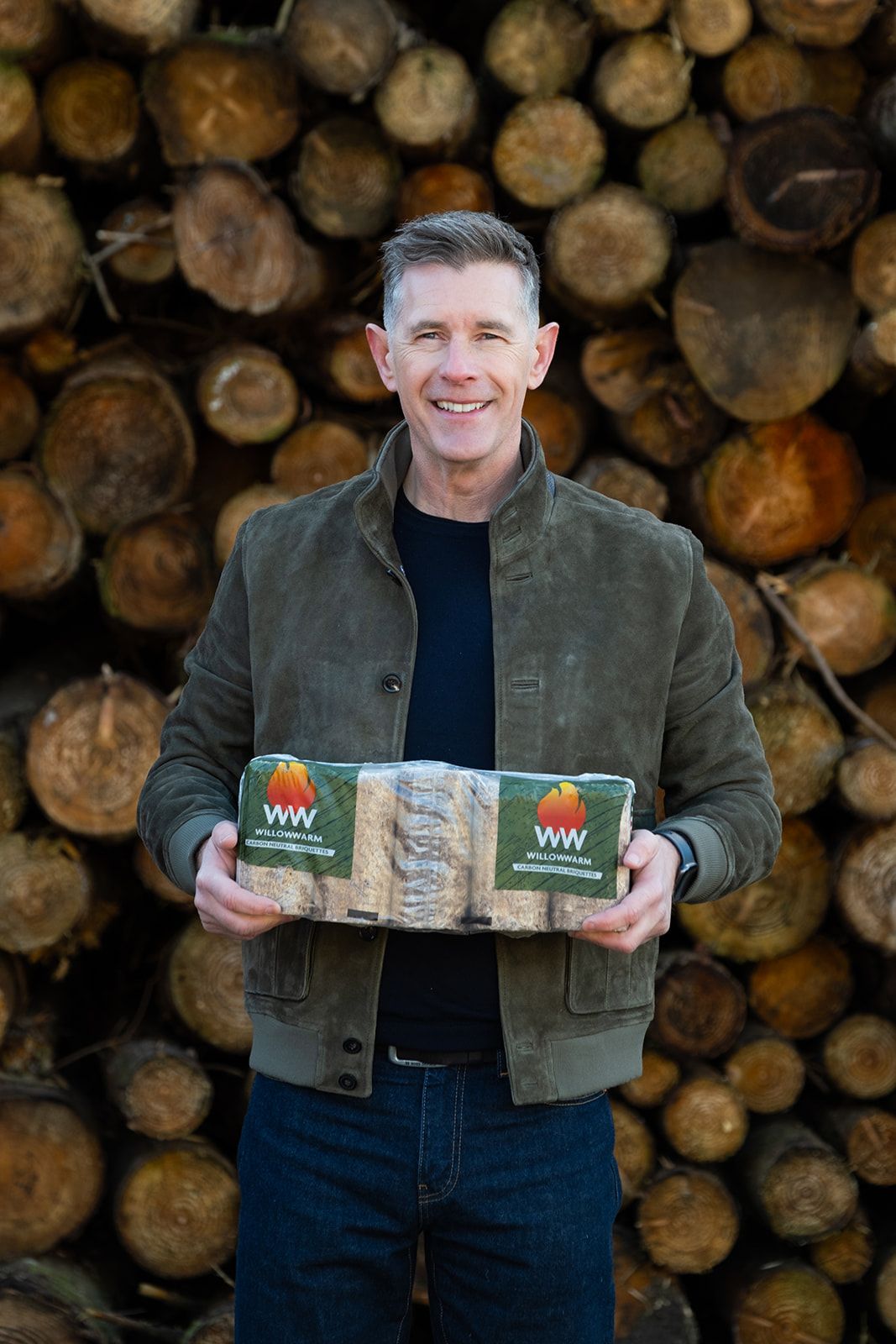 Dermot Bannon smiling holding a bale of WillowWarm Briquettes in front of a lit stove.