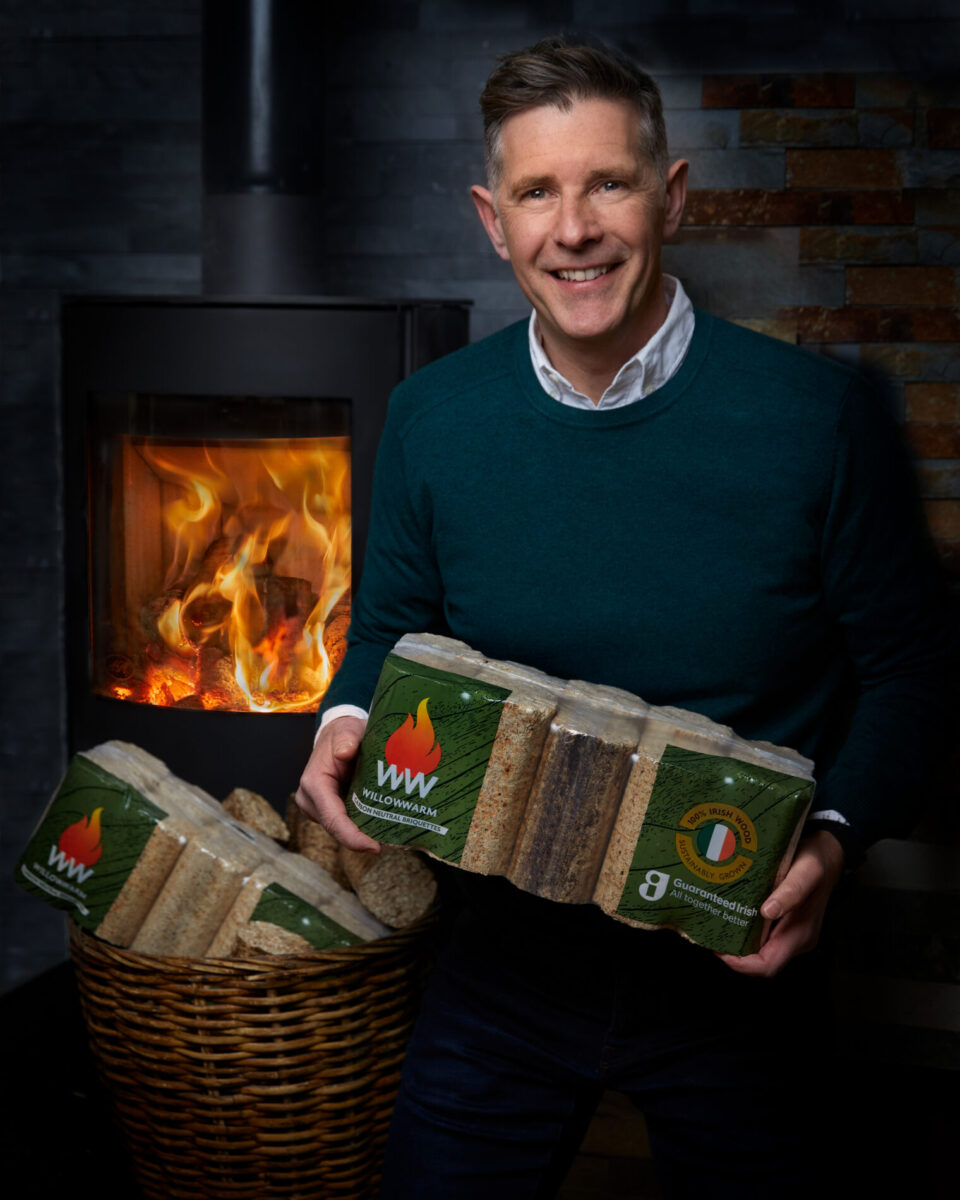 Dermot Bannon smiling holding a bale of WillowWarm Briquettes in front of a lit stove.
