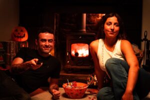 Man and woman sitting in front of a fire, man is holding a television remote control
